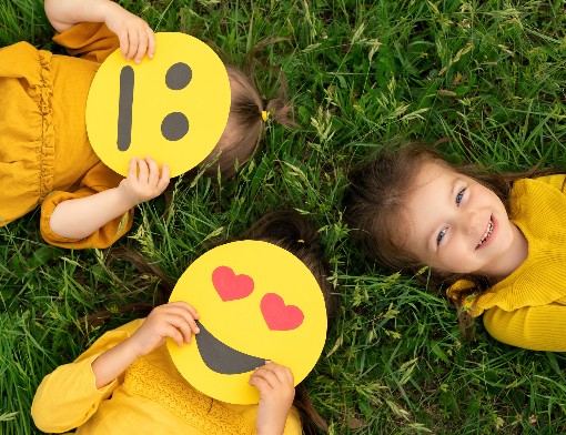 Três crianças deitadas no gramado, duas delas com emojis no rosto
